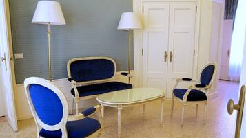 Une pièce comprenant un canapé bleu recouvert de tissu, deux fauteuils, une petite table ronde et deux lampadaires. On y trouve une porte blanche avec une poignée et un interrupteur sur le mur.