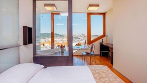 A bedroom with a bed, a desk, and a chair, with a view of a cityscape through large windows. - Wenceslas Square Hotel - Czech Leading Hotels (Prague)
