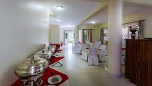 Un salón de banquetes con mesas dispuestas para un evento formal, con manteles blancos y sillas, y una barra de buffet con vajilla de plata.