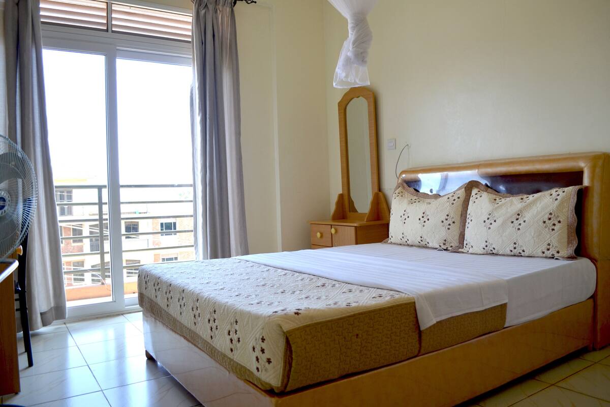 A hotel room with a bed, a wooden nightstand, a mirror, and a window with curtains.
