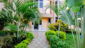 Un patio central con un sendero, rodeado de exuberante vegetación y palmeras, que conduce a un edificio con fachada curva y múltiples ventanas.
