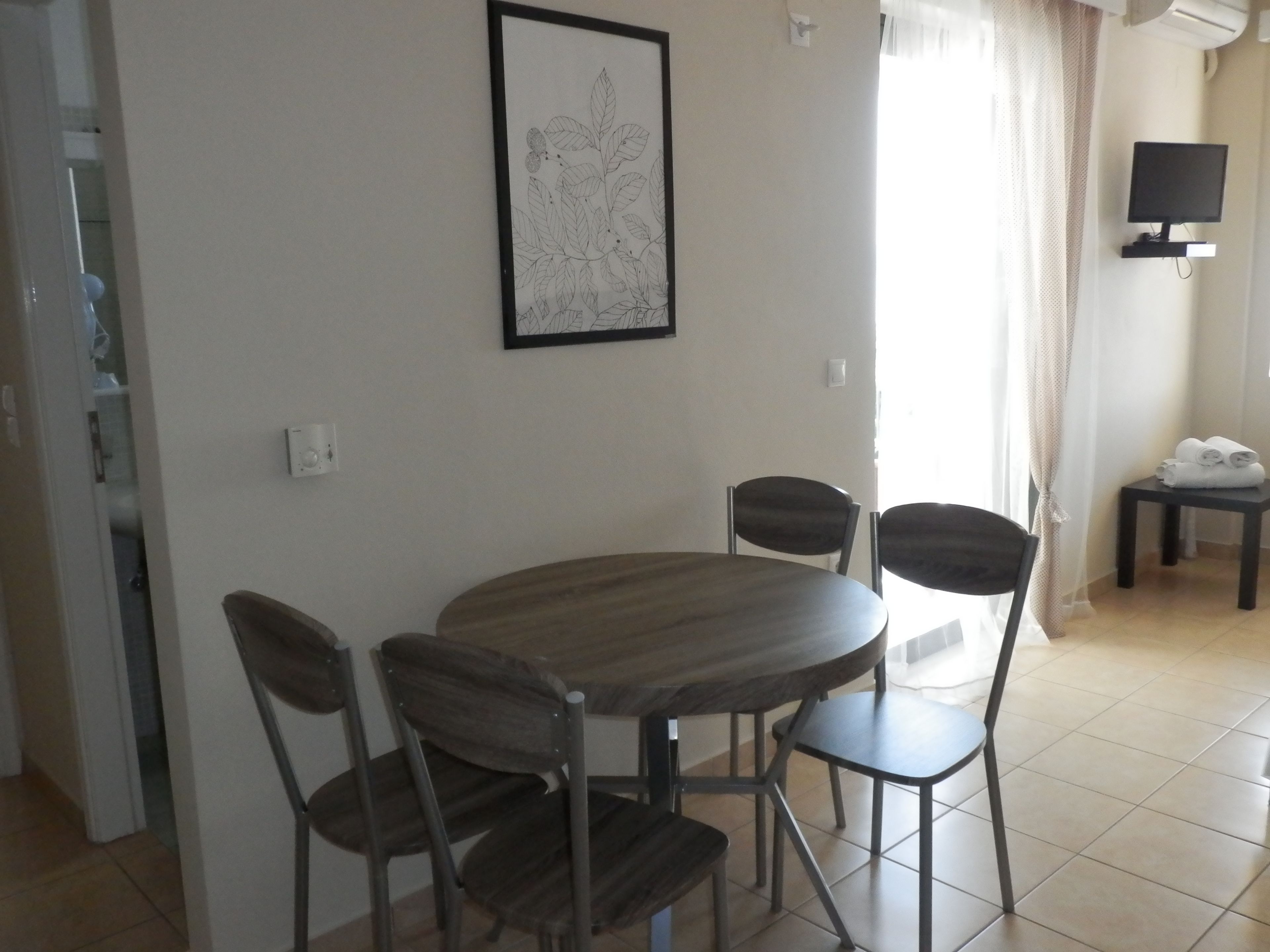 A dining area with a round table and four chairs, a framed botanical print on the wall, a television on a stand, and a small table with towels.