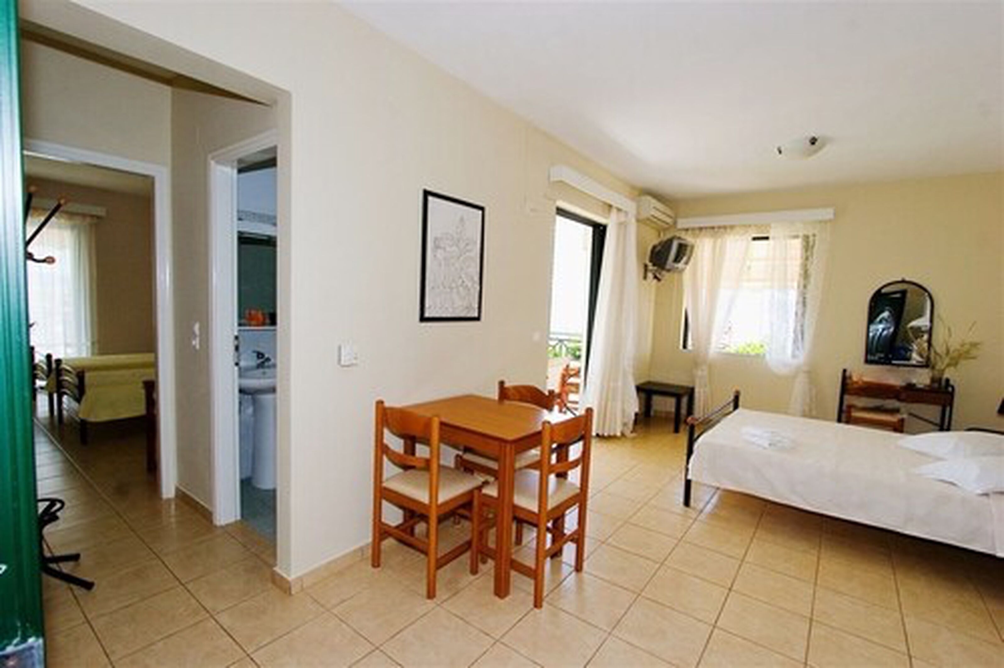 A bedroom with a bed, a small table with chairs, a window with curtains, and a bathroom with a sink and toilet.