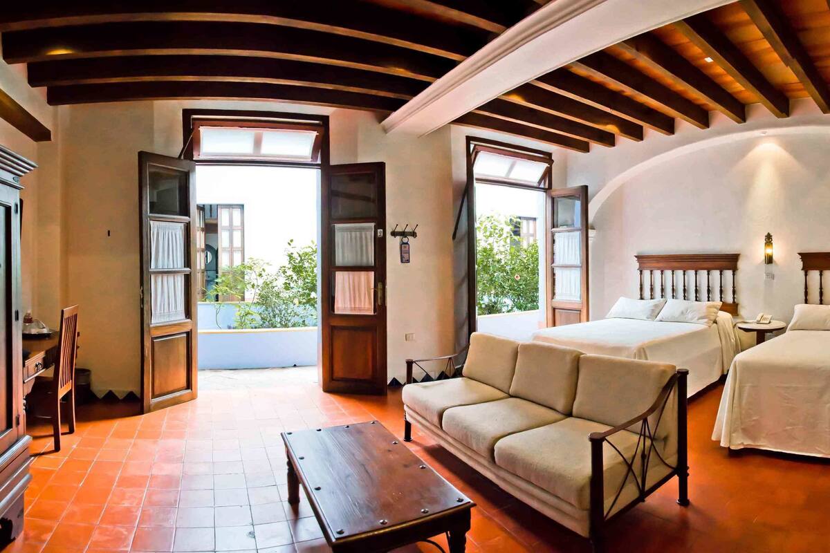 A room with two beds, a sofa, a wooden table, and a view of the outdoors.