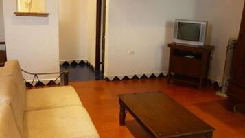 A living room with a sofa, a wooden coffee table, a television on a wooden stand, and a tiled floor.