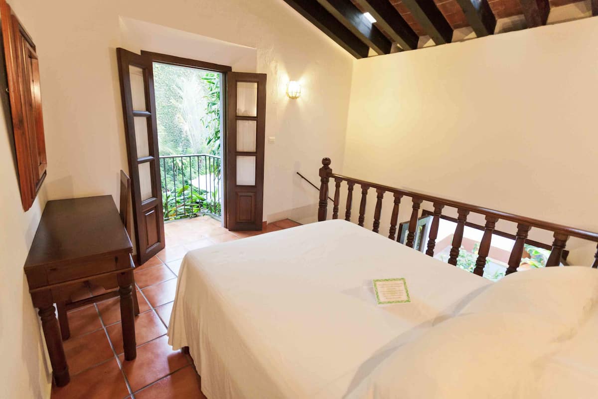 A bedroom with a bed, a wooden desk, and a balcony with a view of greenery.