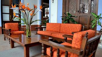 Quarto tradicional com um conjunto de sofá e poltrona de madeira, e uma mesa de centro. Há uma janela grande com cortinas, uma porta de madeira com detalhes entalhados e um vaso com planta.