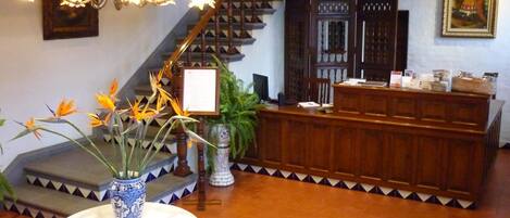 A reception area with a wooden desk, a vase with flowers, a staircase, and a chandelier.
