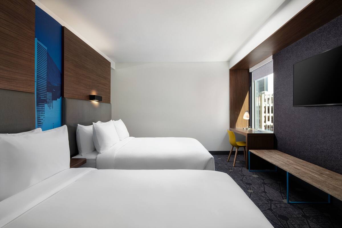 Habitación de hotel con dos camas, un escritorio, una silla, un banco, un televisor y una ventana con vista a la ciudad.
