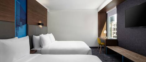 A hotel room with two beds, a desk, a chair, a TV, and a window.