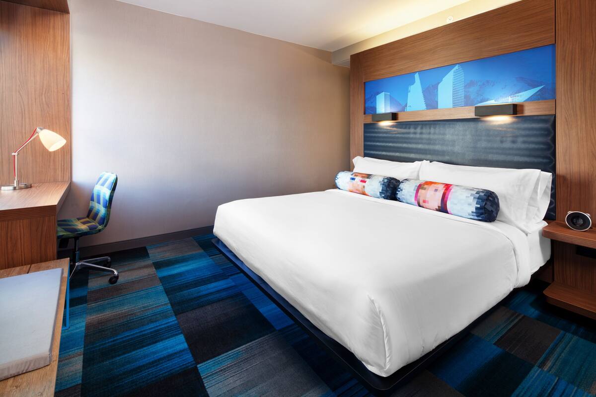 Habitación de hotel con cama, escritorio con lámpara, una silla y una obra de arte con vista a la ciudad.