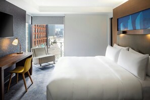 Habitación de hotel con cama, escritorio, silla, televisor y vista a la ciudad.