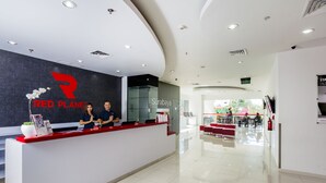 Una recepción con logotipo, dos empleados, un separador de vidrio y un interior moderno con techo curvo y un área de comedor visible al fondo.