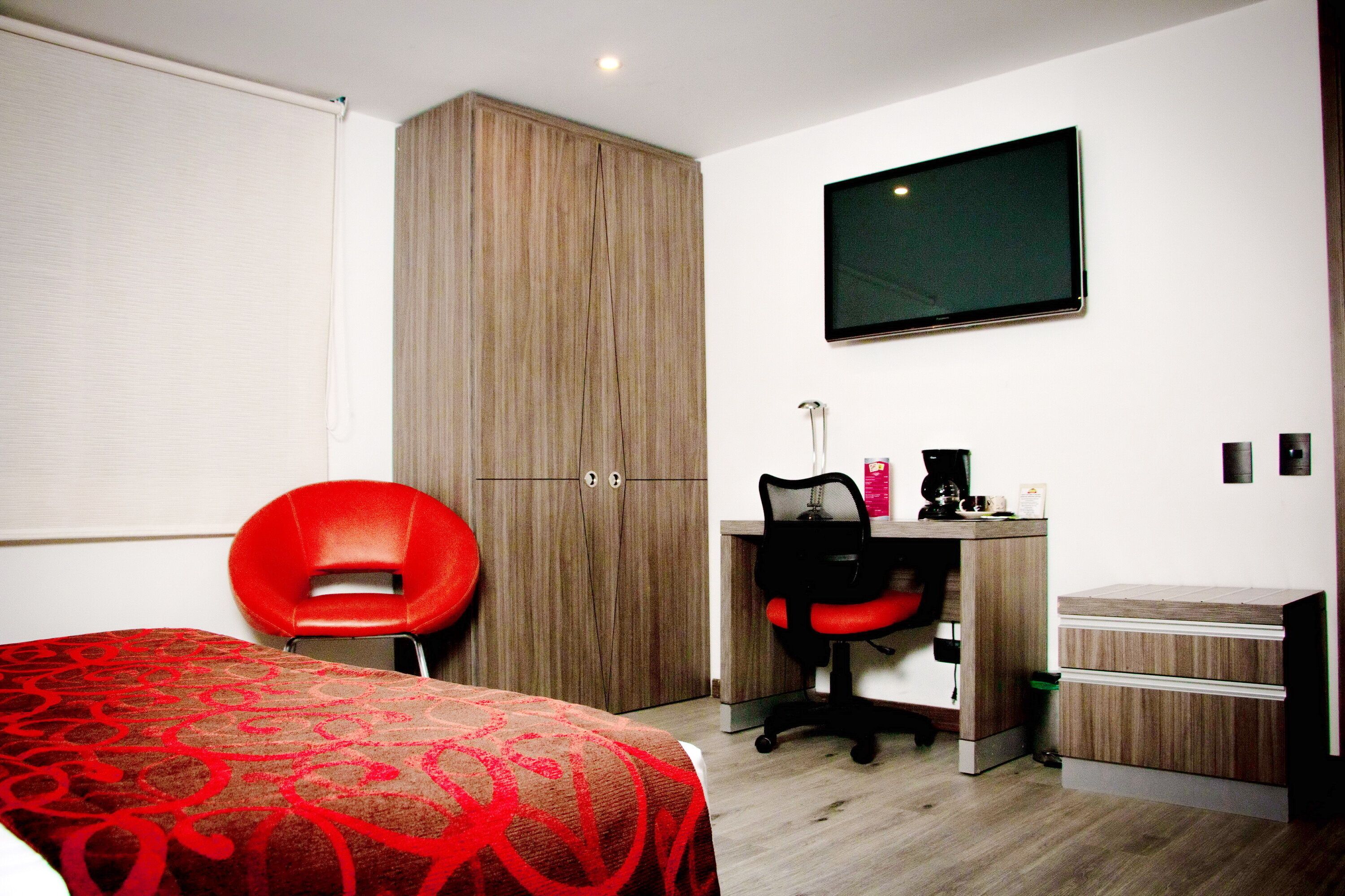 Quarto de hotel com uma cama, uma cadeira vermelha, uma mesa com um computador e uma televisão na parede.
