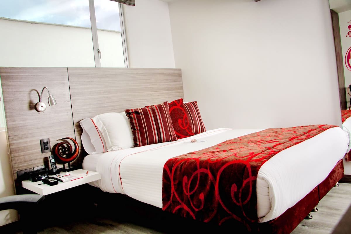 A hotel room with a bed, a bedside table with a lamp, a telephone, and a red and white patterned bedspread.