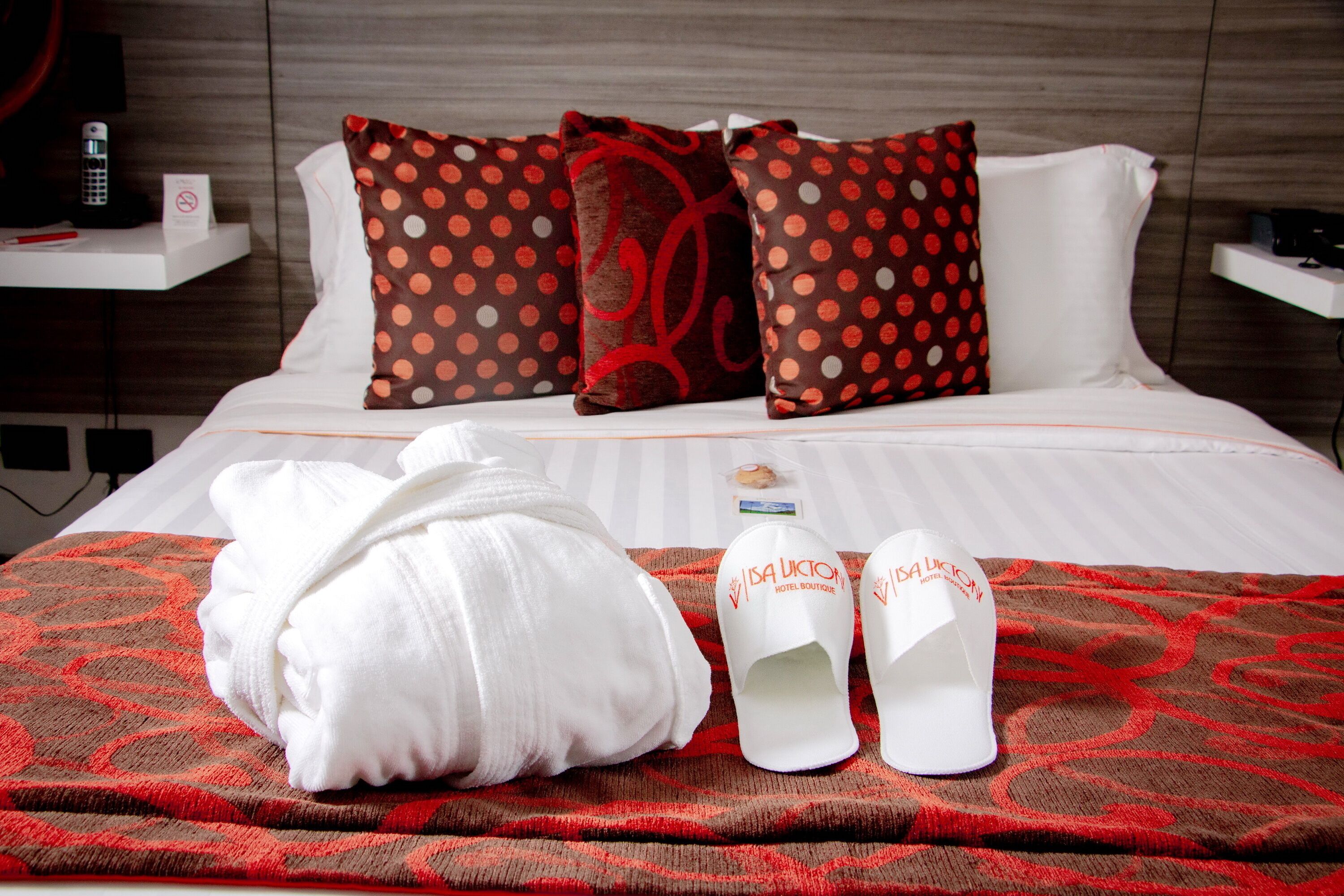 A neatly made bed with a red and brown patterned blanket, white pillows with red polka dots, a white bathrobe, and a pair of white slippers with a logo.