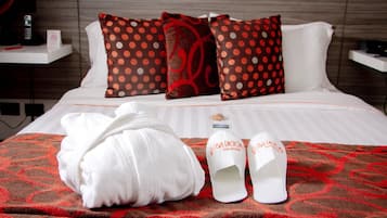 A neatly made bed with a red and brown patterned blanket, white pillows with red polka dots, a white bathrobe, and a pair of white slippers with a logo.