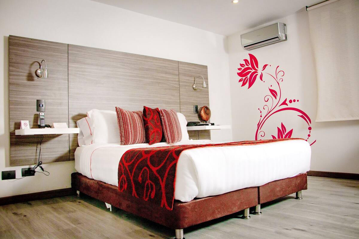 A modern hotel room with a wooden headboard, a bed with white bedding and red accents, a bedside table with a lamp, and a decorative wall sticker.