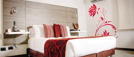 A modern hotel room with a wooden headboard, a bed with white bedding and red accents, a bedside table with a lamp, and a decorative wall sticker.