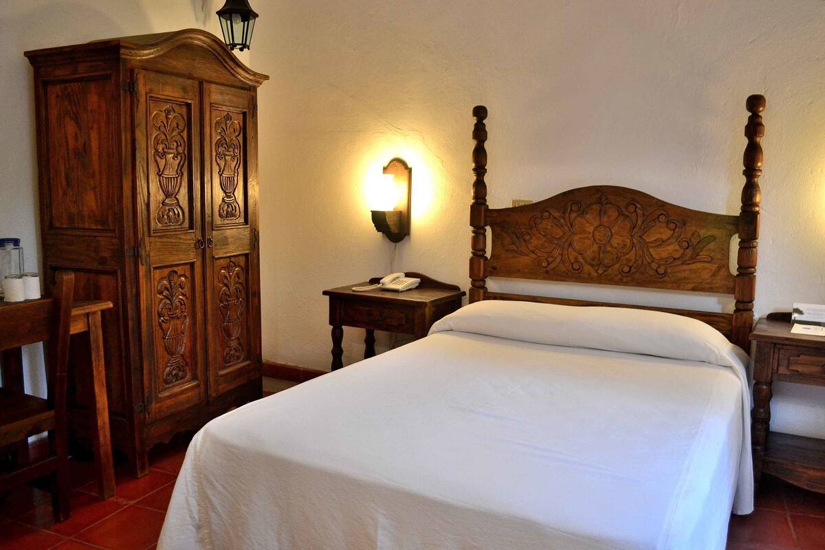 Una cama bien hecha con cabecera de madera, un armario de madera y una mesita de noche con lámpara.