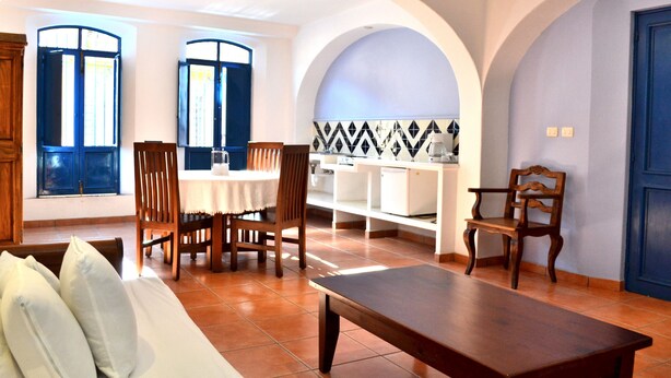 Un área de comedor con una mesa y sillas, un sofá y un banco. Hay dos ventanas con contraventanas azules y un salpicadero de azulejos.
