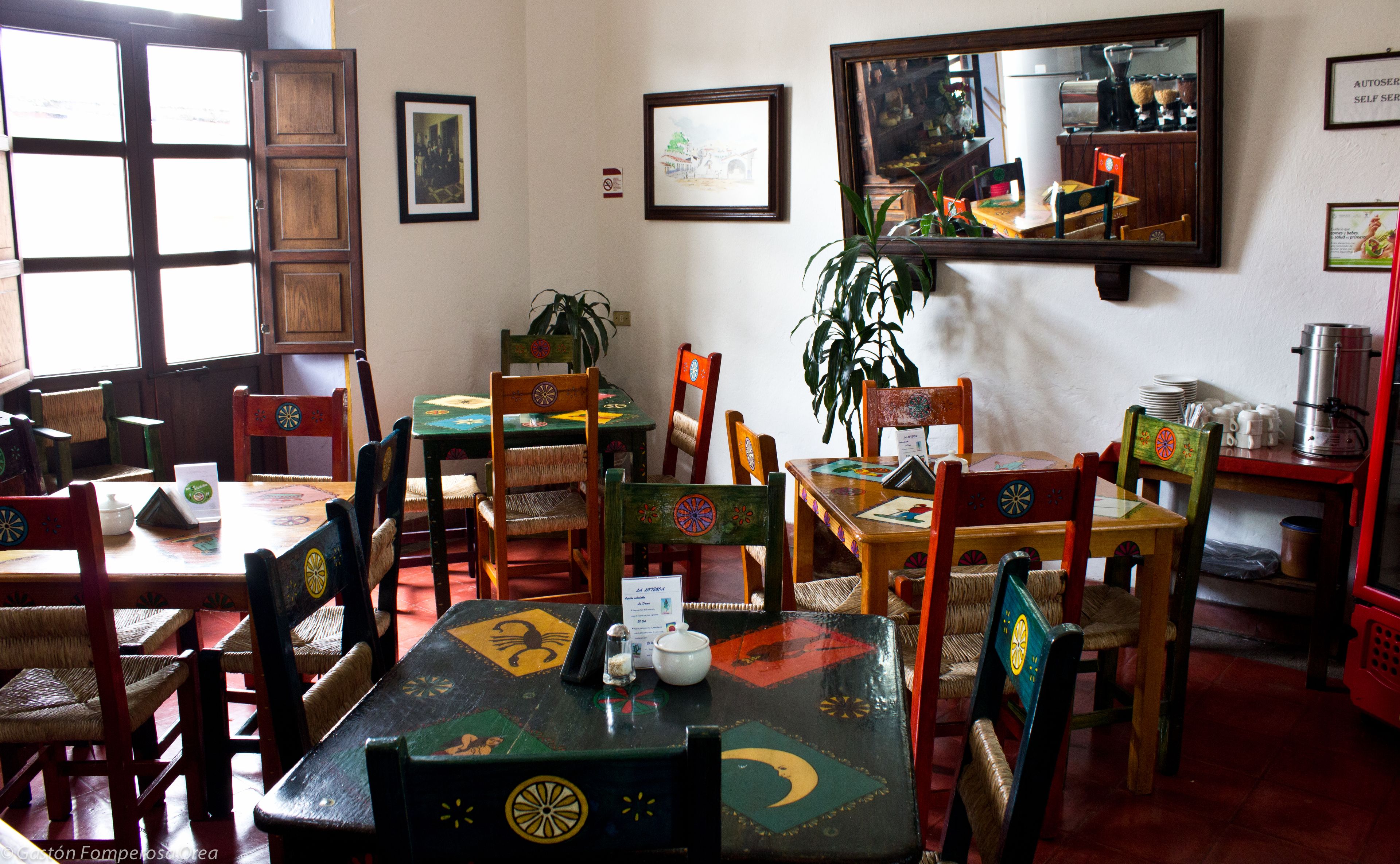 A restaurant interior with wooden tables and chairs, framed pictures on the walls, and a large mirror reflecting the room.