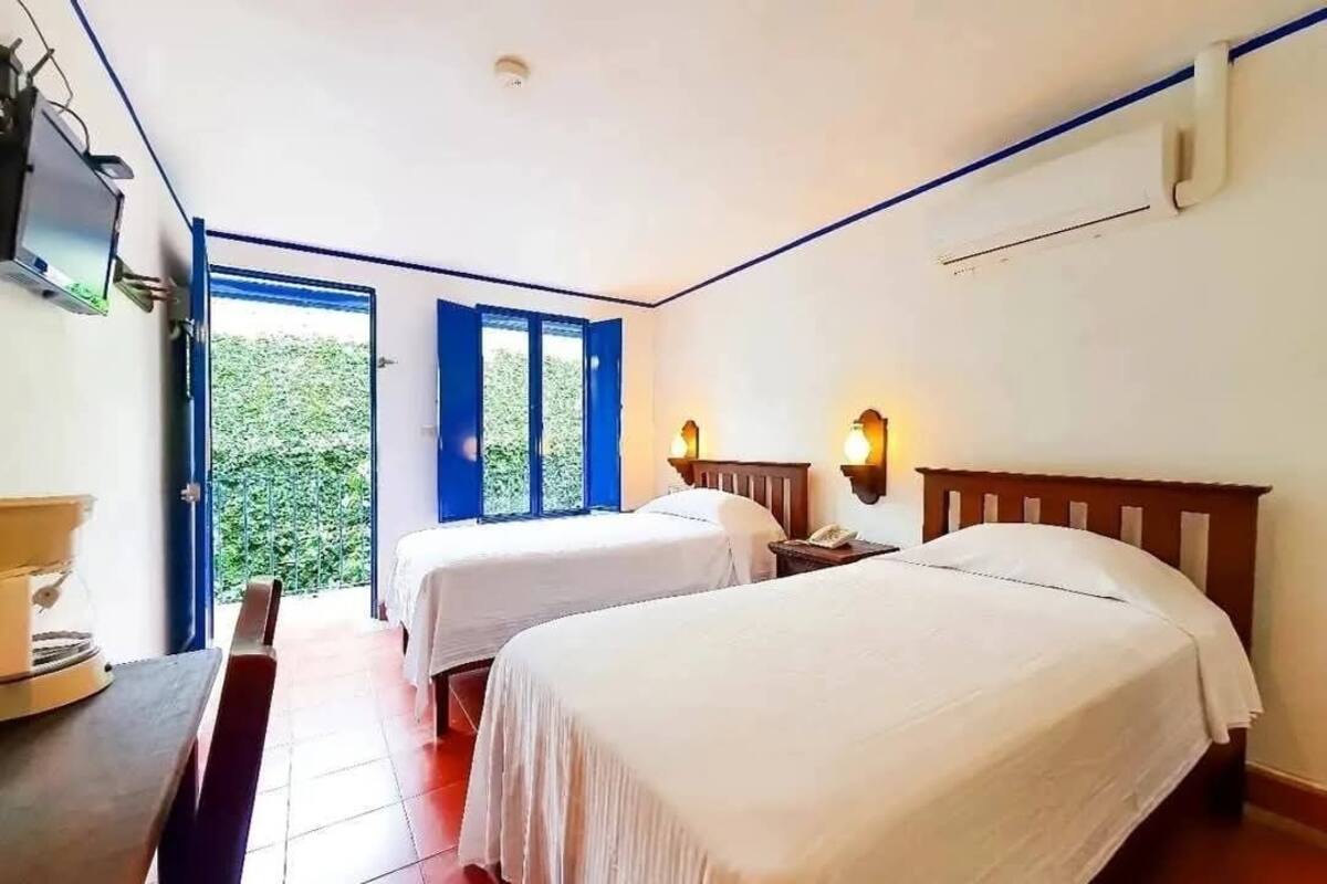 Habitación de hotel con dos camas, un escritorio y vistas a la vegetación desde la puerta.