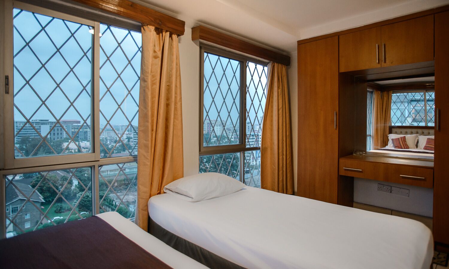 A hotel room with two beds, a wooden wardrobe, and large windows with curtains.
