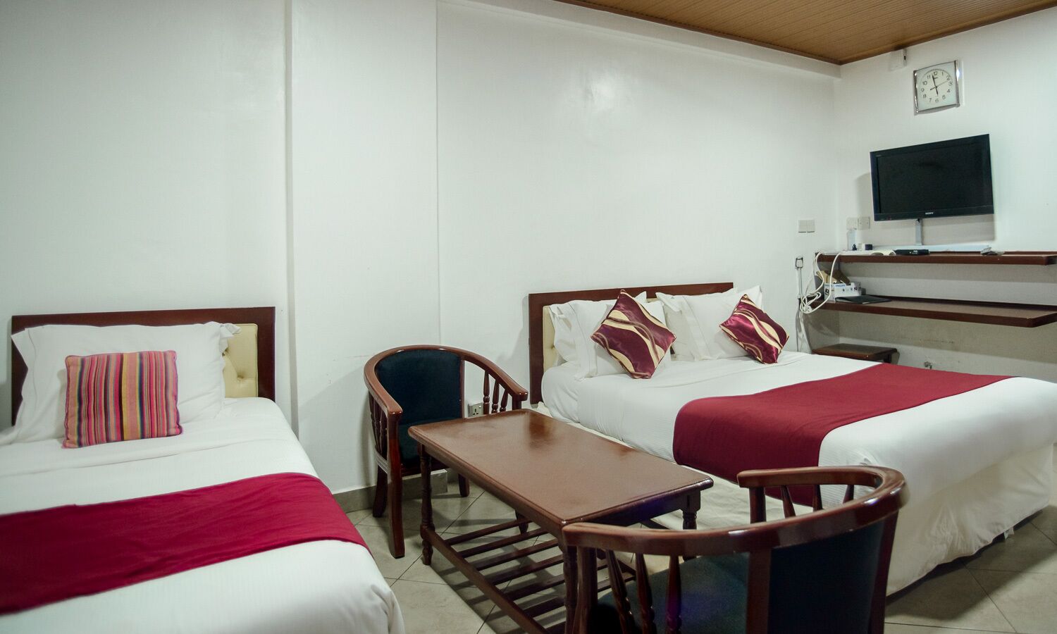 A hotel room with two beds, a desk, a chair, a TV, and a clock on the wall.