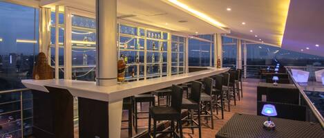 Zona de bar moderna con cerramiento de vidrio, taburetes y vistas de la ciudad.