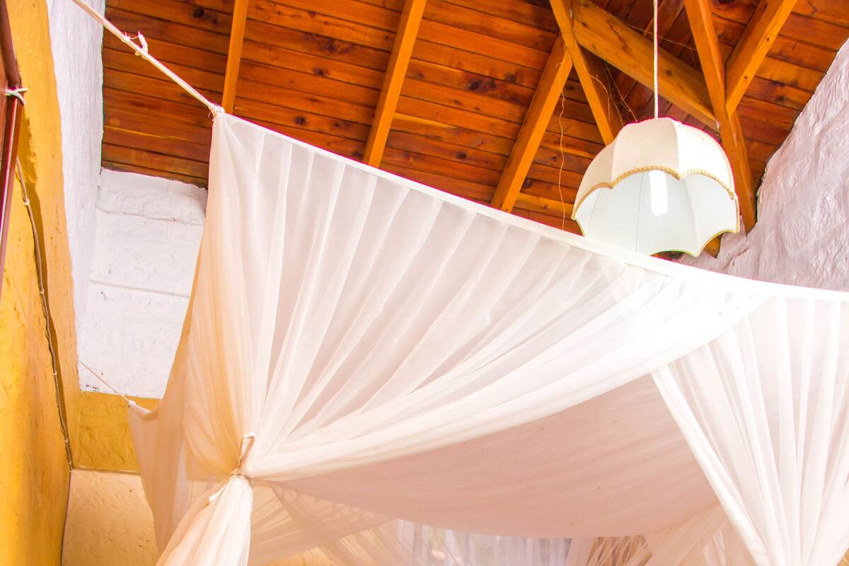 A room with a bed under a canopy, wooden ceiling beams, a window with curtains, and a bedside table.