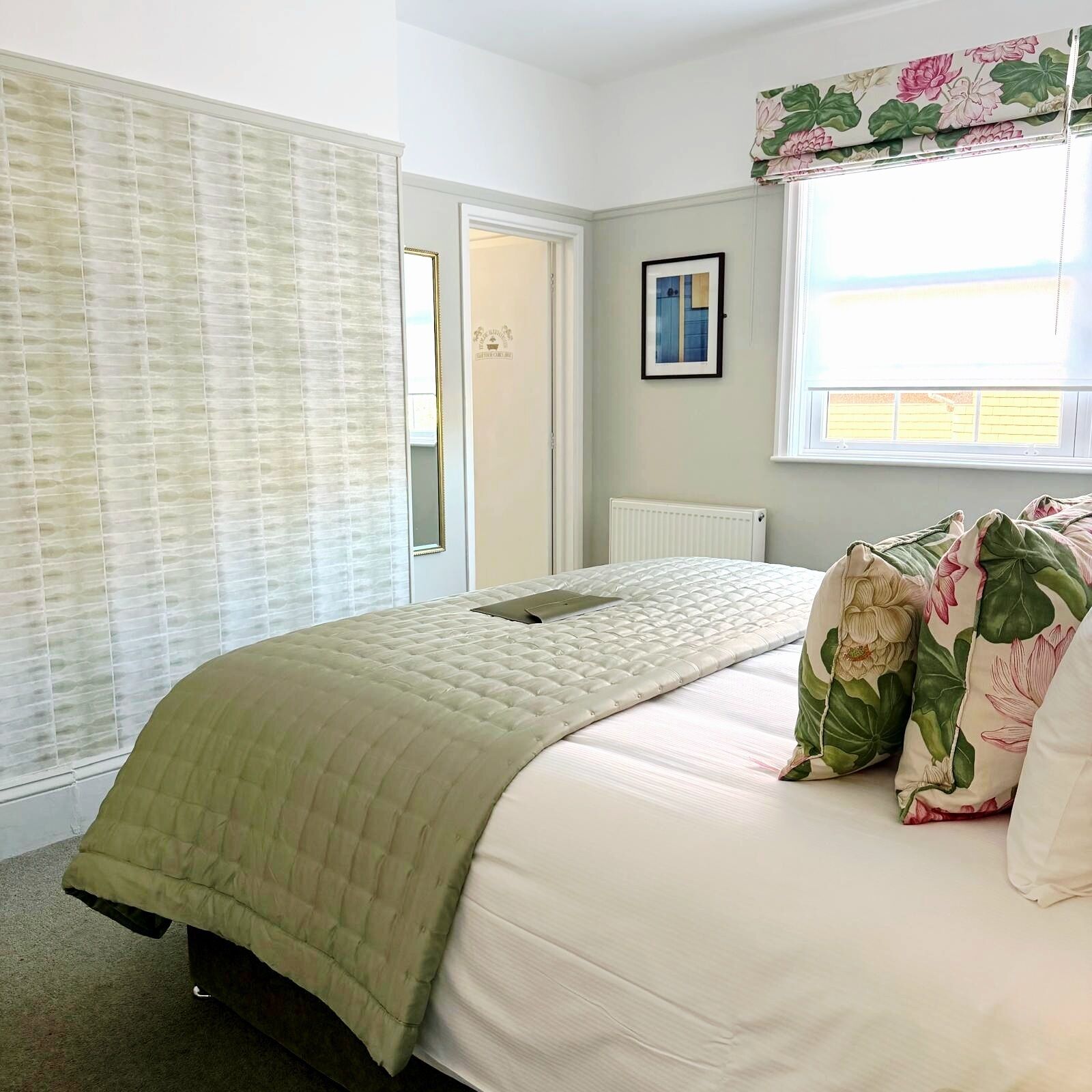 A bedroom with a bed, a radiator, a window with floral curtains, and a door.
