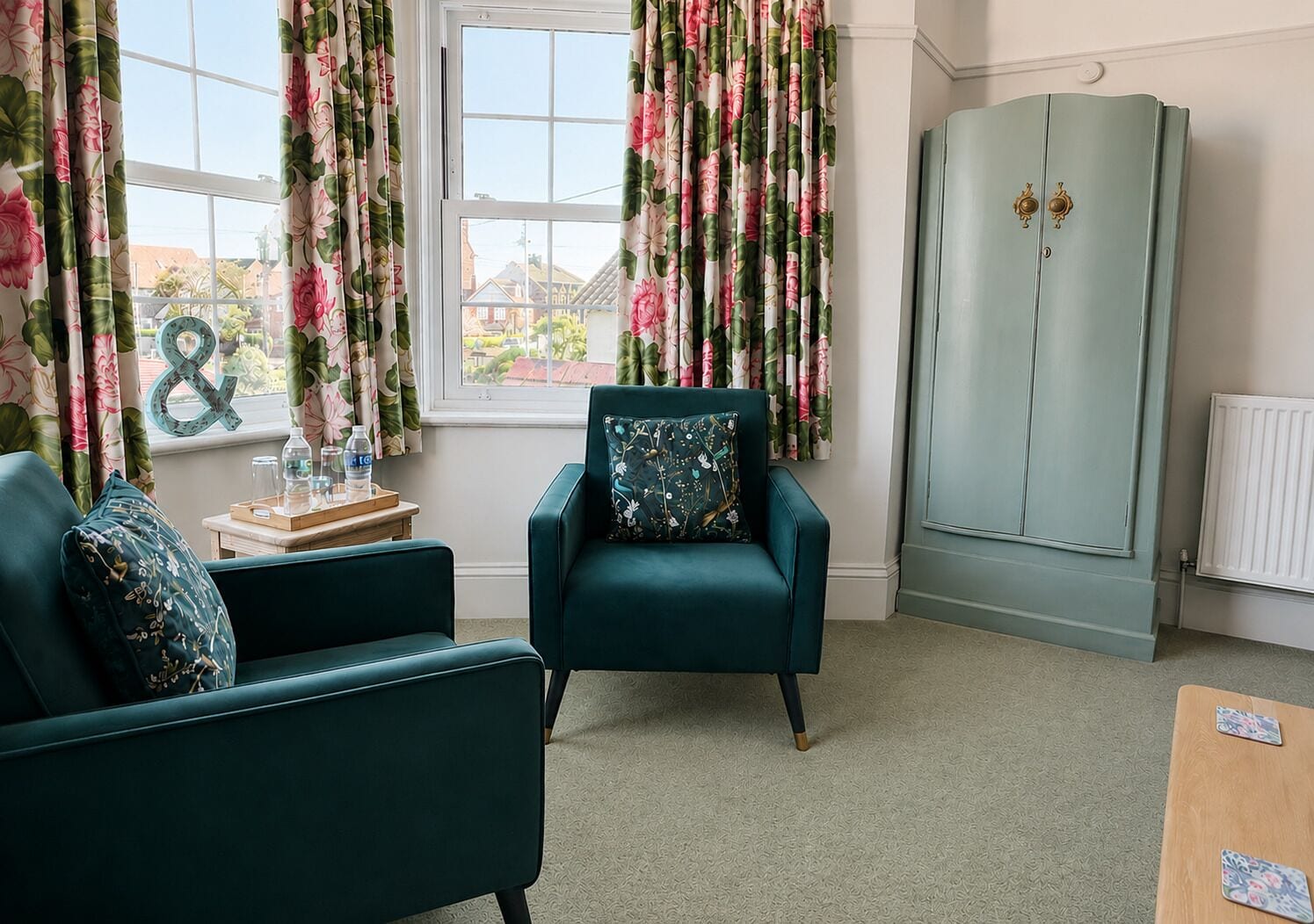 A room with teal armchairs, a wooden side table with water bottles, a radiator, and a wardrobe.