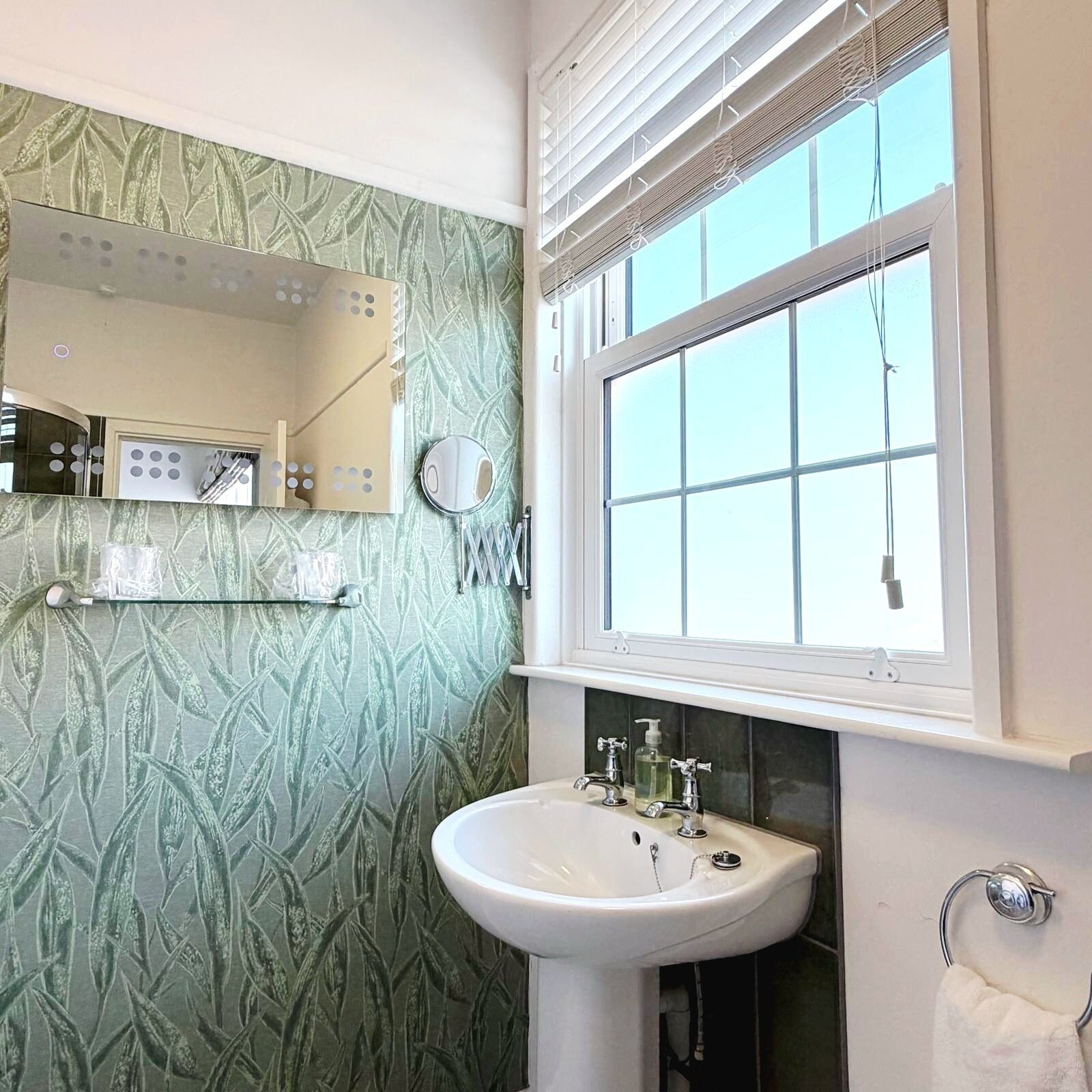 Baño con lavamanos, espejo, ventana y papel tapiz estampado.