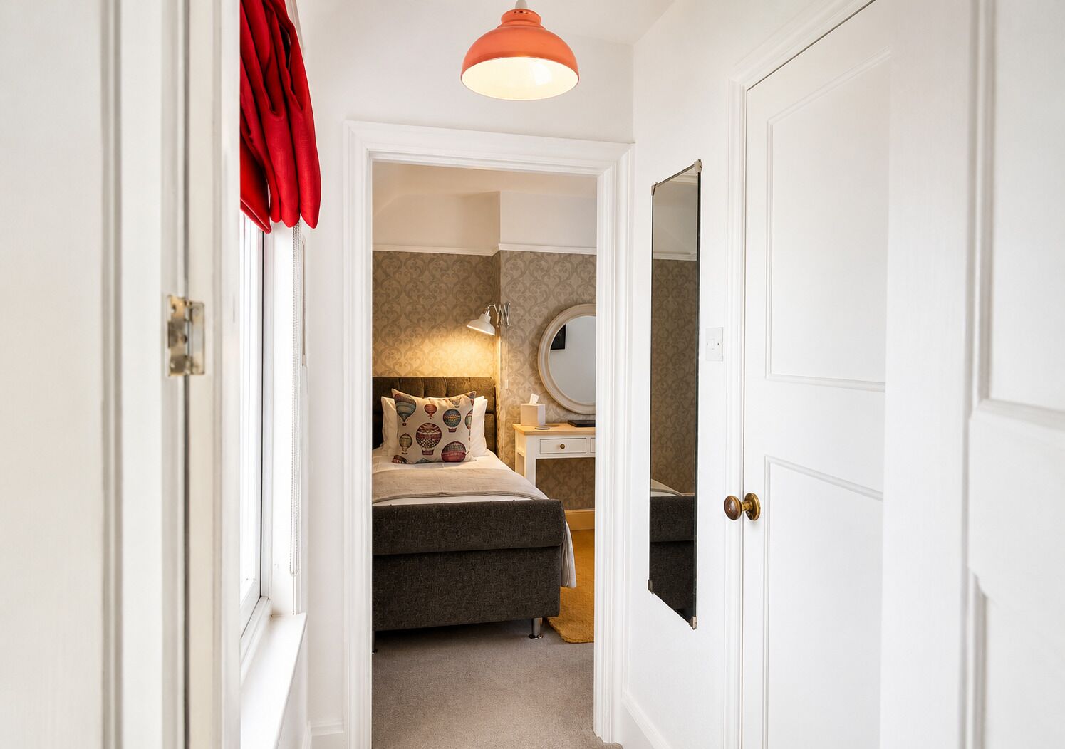A narrow hallway leading to a bedroom with a bed, bedside table, and a mirror.