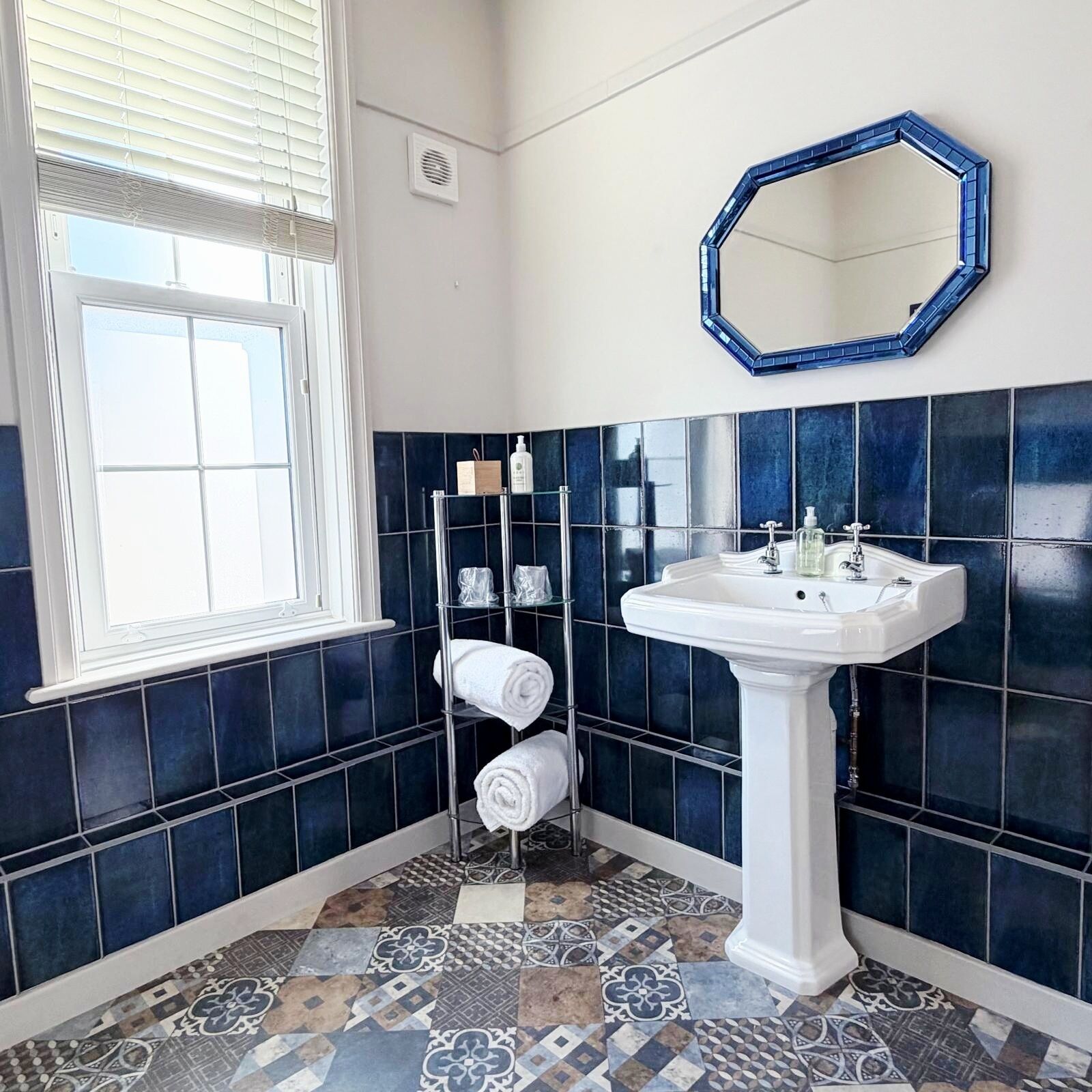 A bathroom with dark blue tiles, a white sink, a mirror, and a window with blinds.