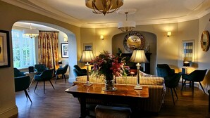 A spacious living room with a wooden coffee table, a sofa, and multiple chairs. There is a fireplace with a mirror above it, and a chandelier hanging from the ceiling. - Burlington Berties (Sheringham)