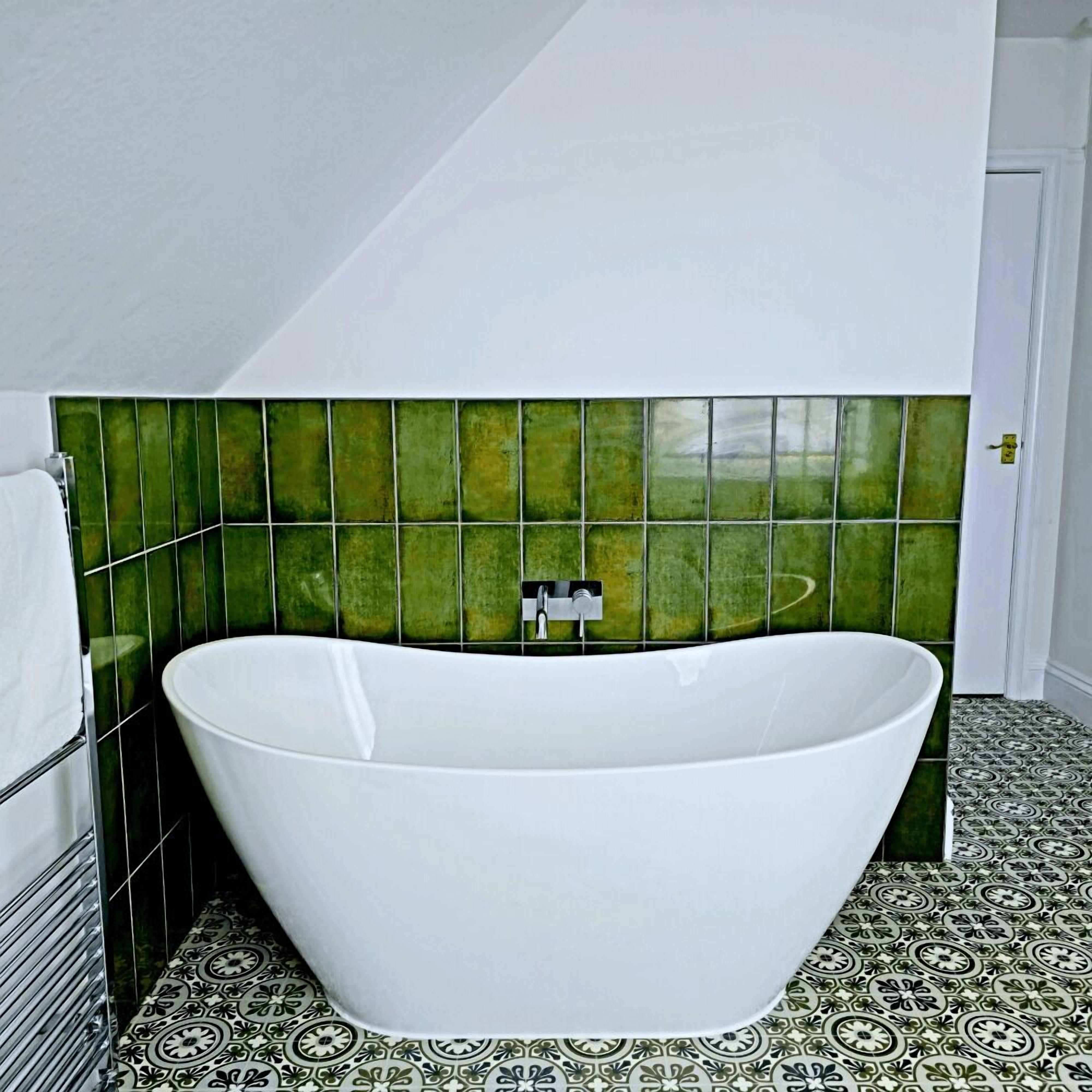 A modern bathroom with a freestanding bathtub, green tiled walls, and patterned floor tiles.