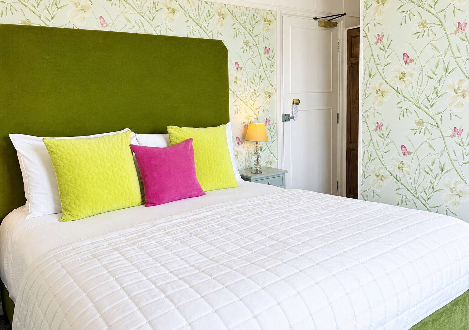 Un lit avec une tête de lit verte, des oreillers blancs et roses, et un papier peint à motifs floraux.