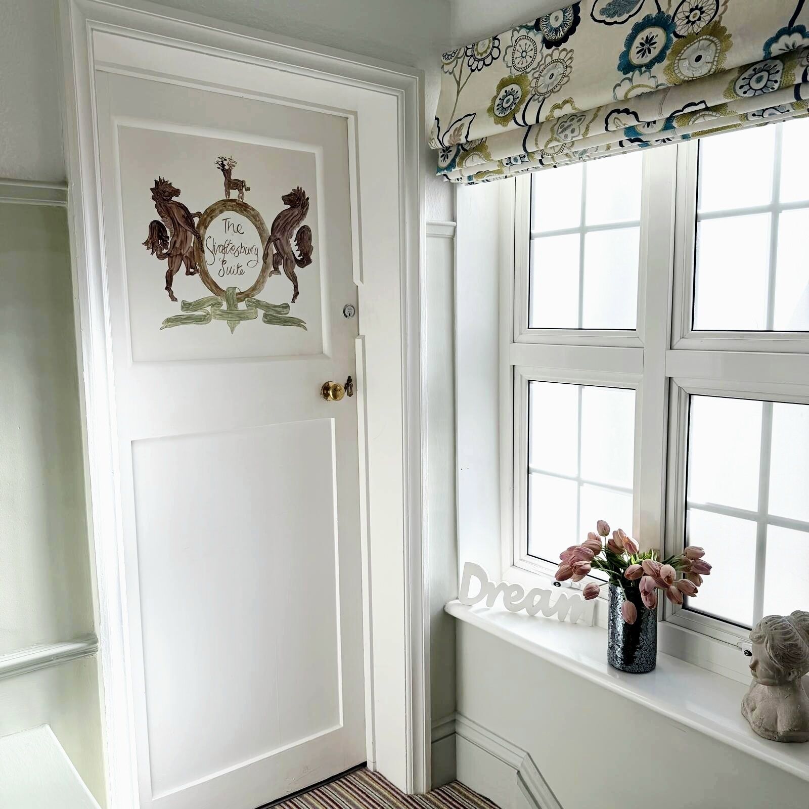 A white door with a decorative plaque, a window with a view, and a vase with flowers on the windowsill.