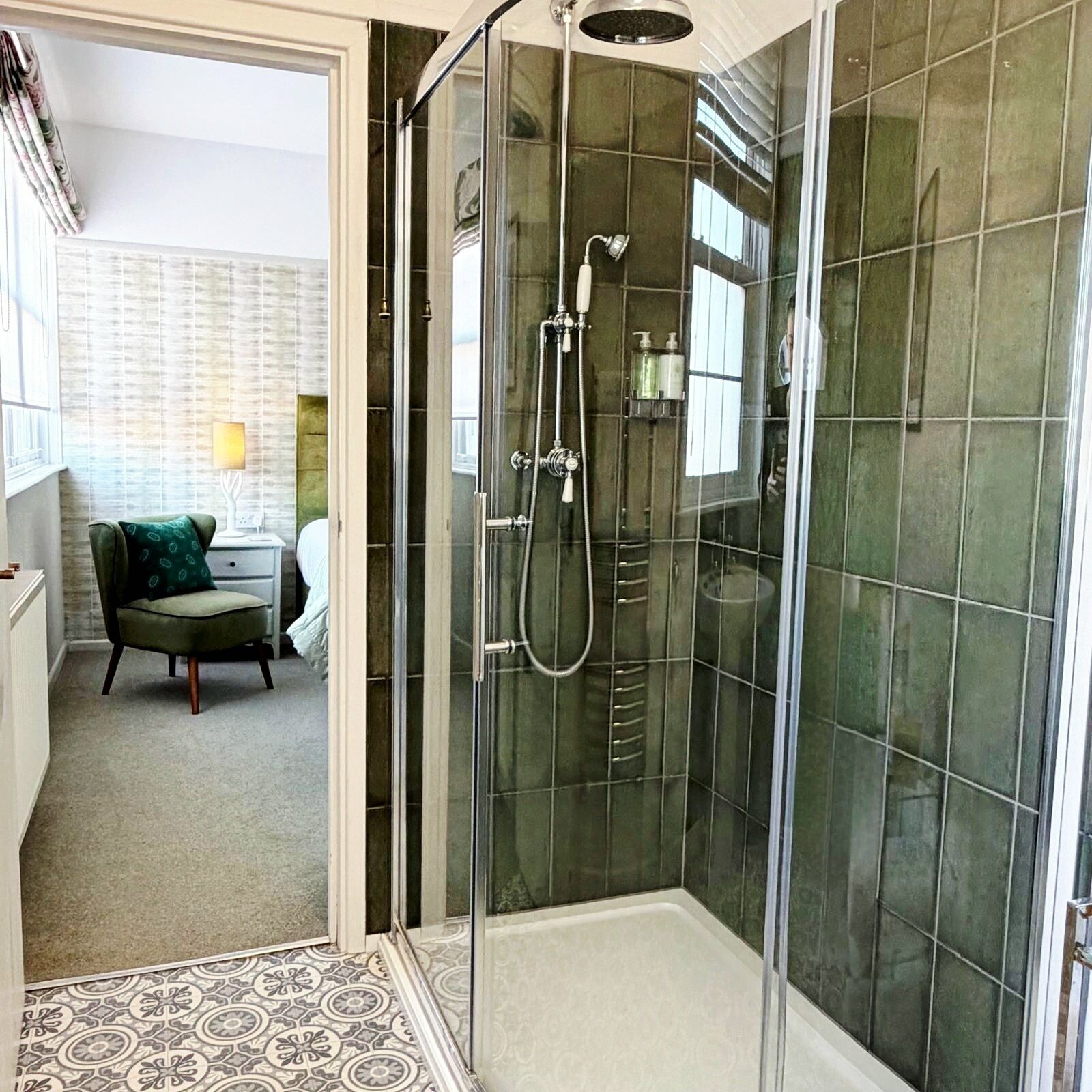 A modern bathroom with a glass-enclosed shower, a patterned floor, and a view of a living area with a chair and lamp.