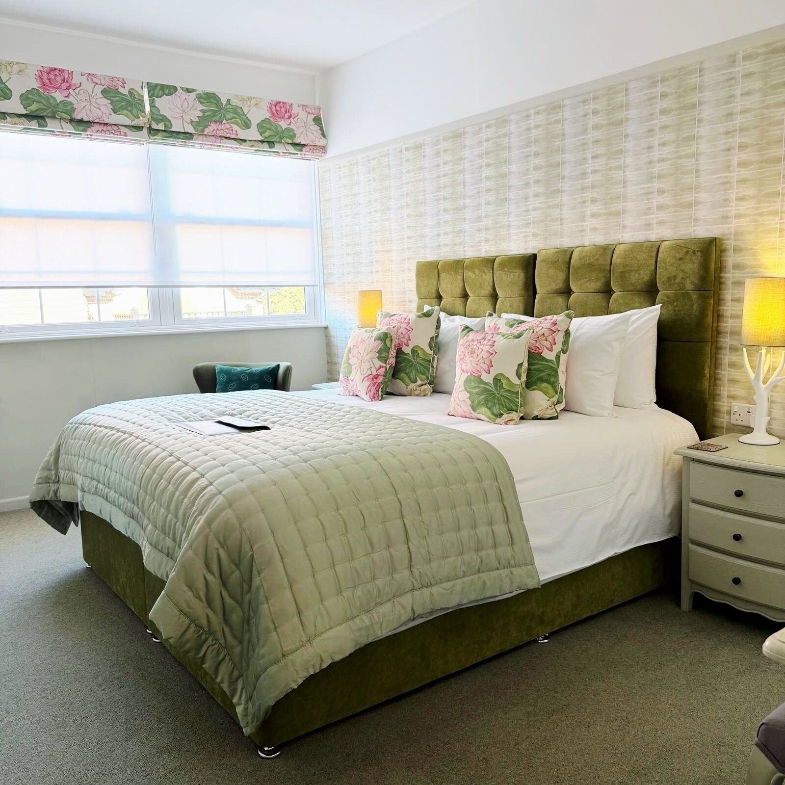 A bedroom with a green upholstered bed, floral-patterned pillows, a bedside table with a lamp, and a window with a floral curtain.