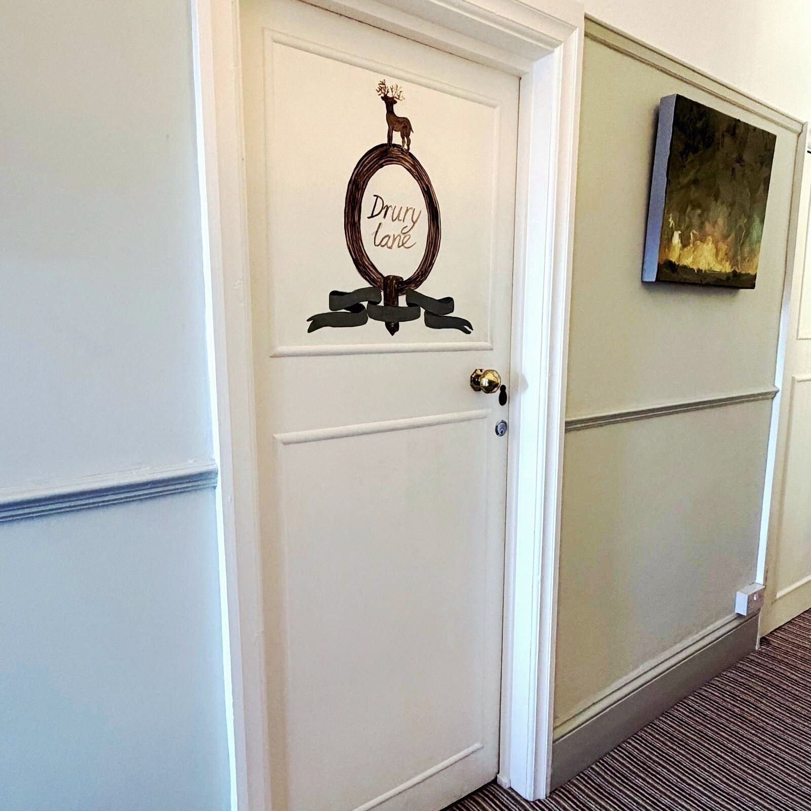 A white door with a deer head and 'Drury Lane' sign, a brass doorknob, and a framed artwork on the adjacent wall.