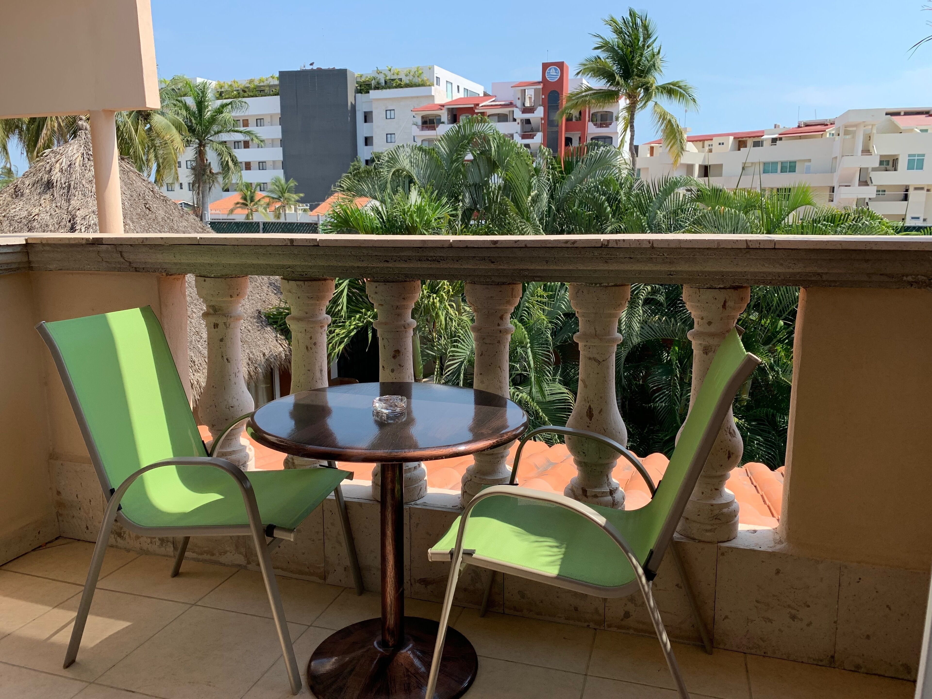 A balcony with a round table and two chairs, overlooking a residential area with buildings and palm trees.