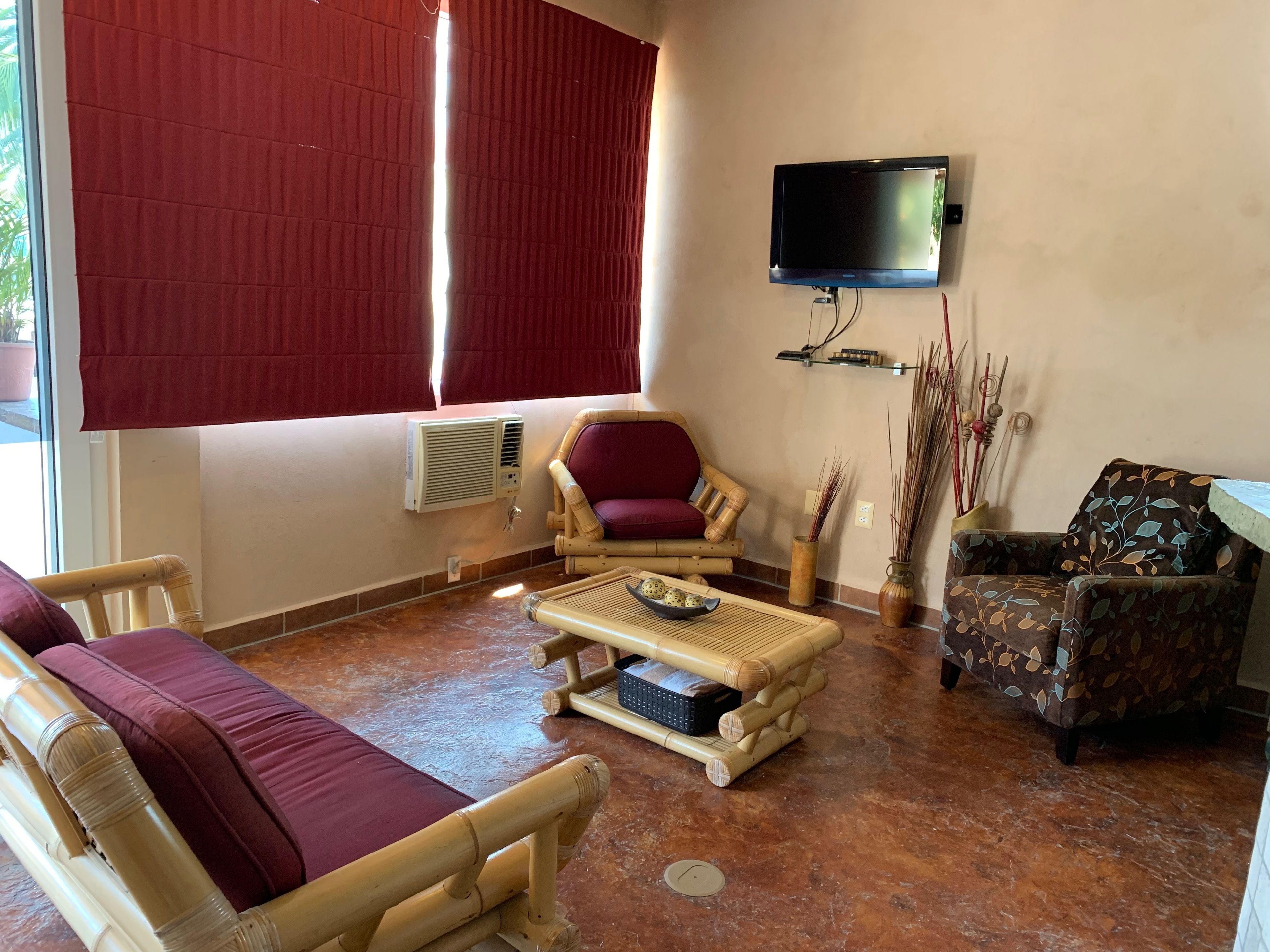 A room with a TV mounted on the wall, a large window with red curtains, a bamboo chair, a cushioned sofa, and a patterned armchair.