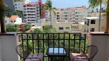 Un balcon avec deux chaises et une table, d’où l’on peut admirer un paysage urbain avec des immeubles et des palmiers.