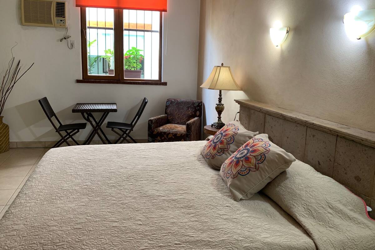Une chambre à coucher comprenant un lit, deux chaises, une petite table, une lampe et une fenêtre avec des stores.