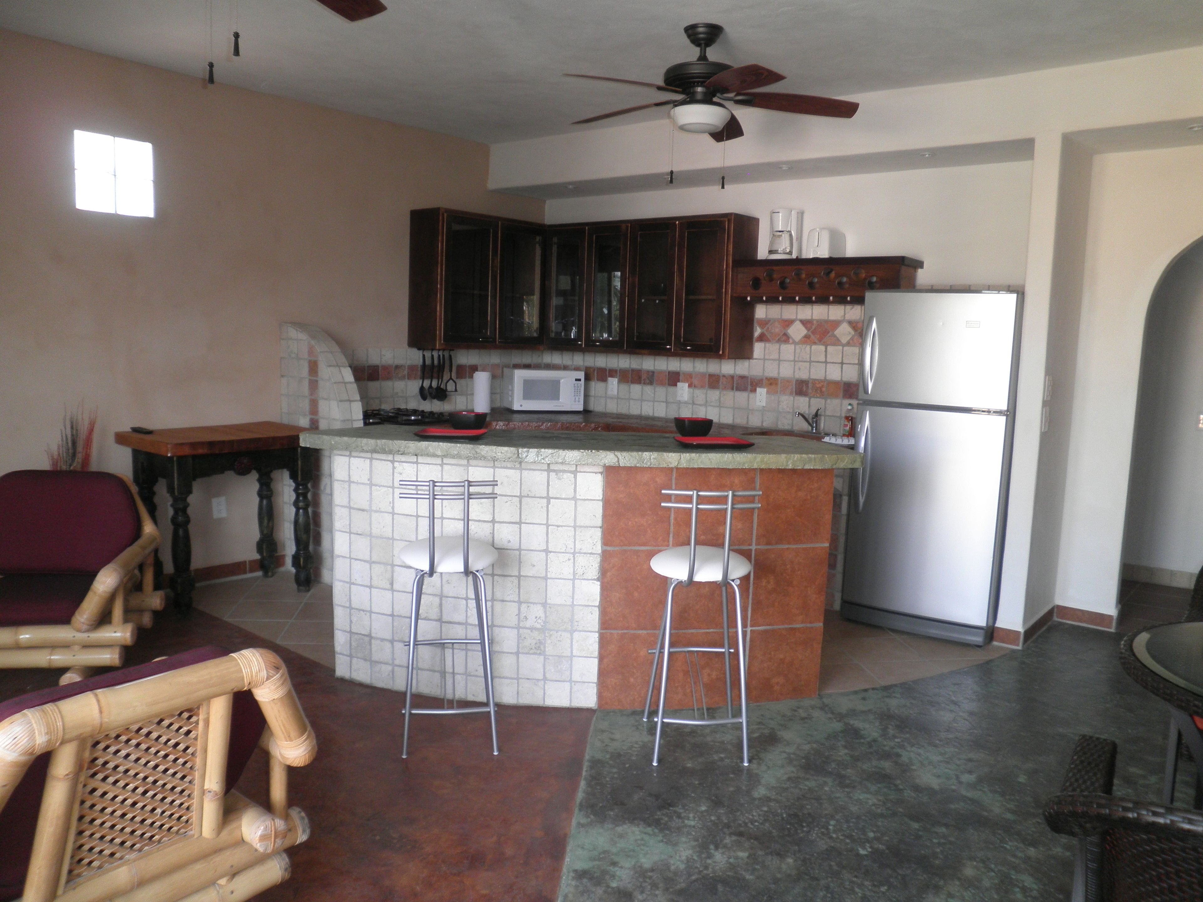 Una cucina compatta con isola centrale, sgabelli da bar, forno a microonde, frigorifero e ventilatore a soffitto.