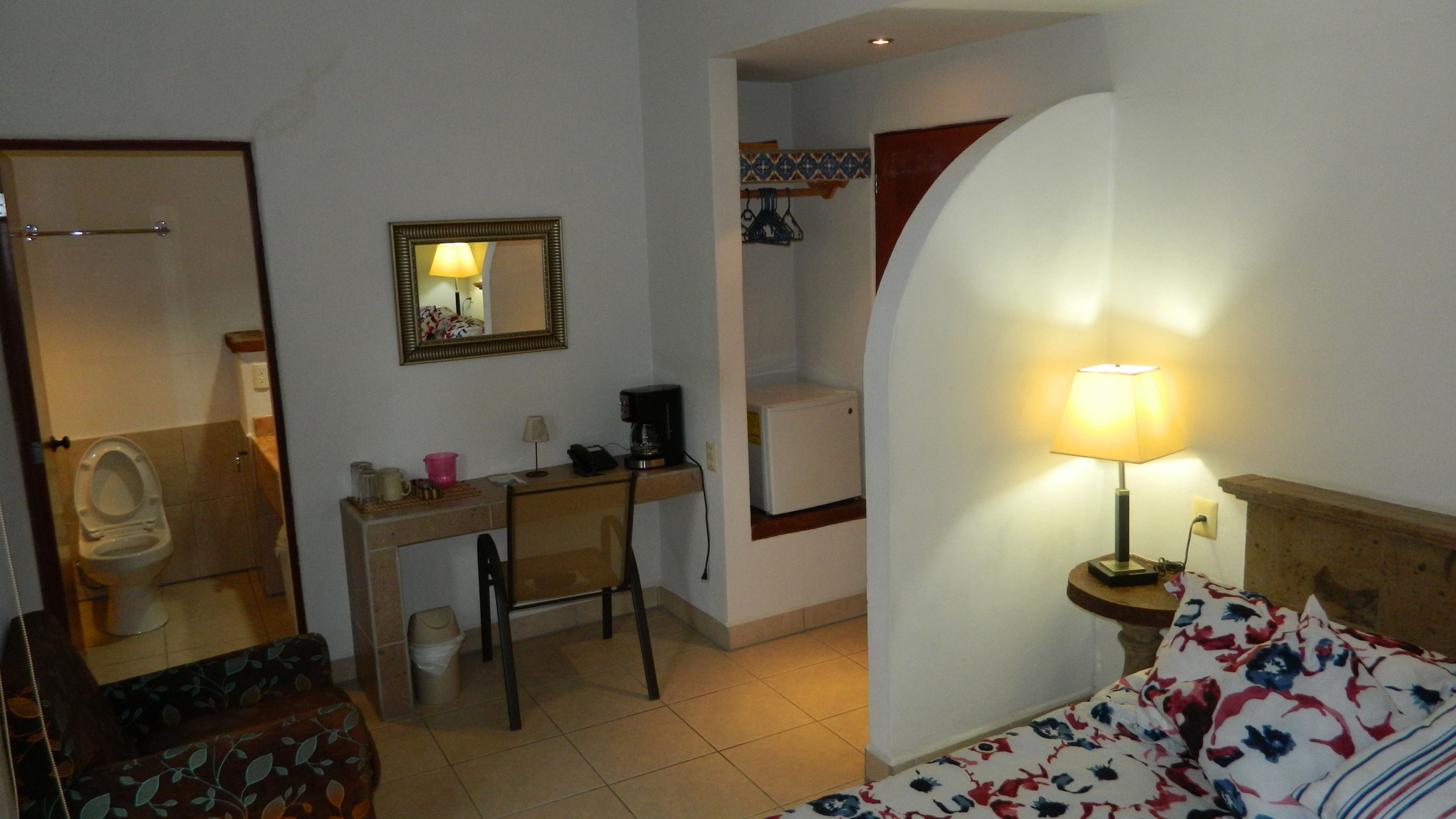 Une chambre d’hôtel comprenant un lit, un bureau avec une chaise, un petit réfrigérateur, une machine à café, une lampe, un miroir et des toilettes.