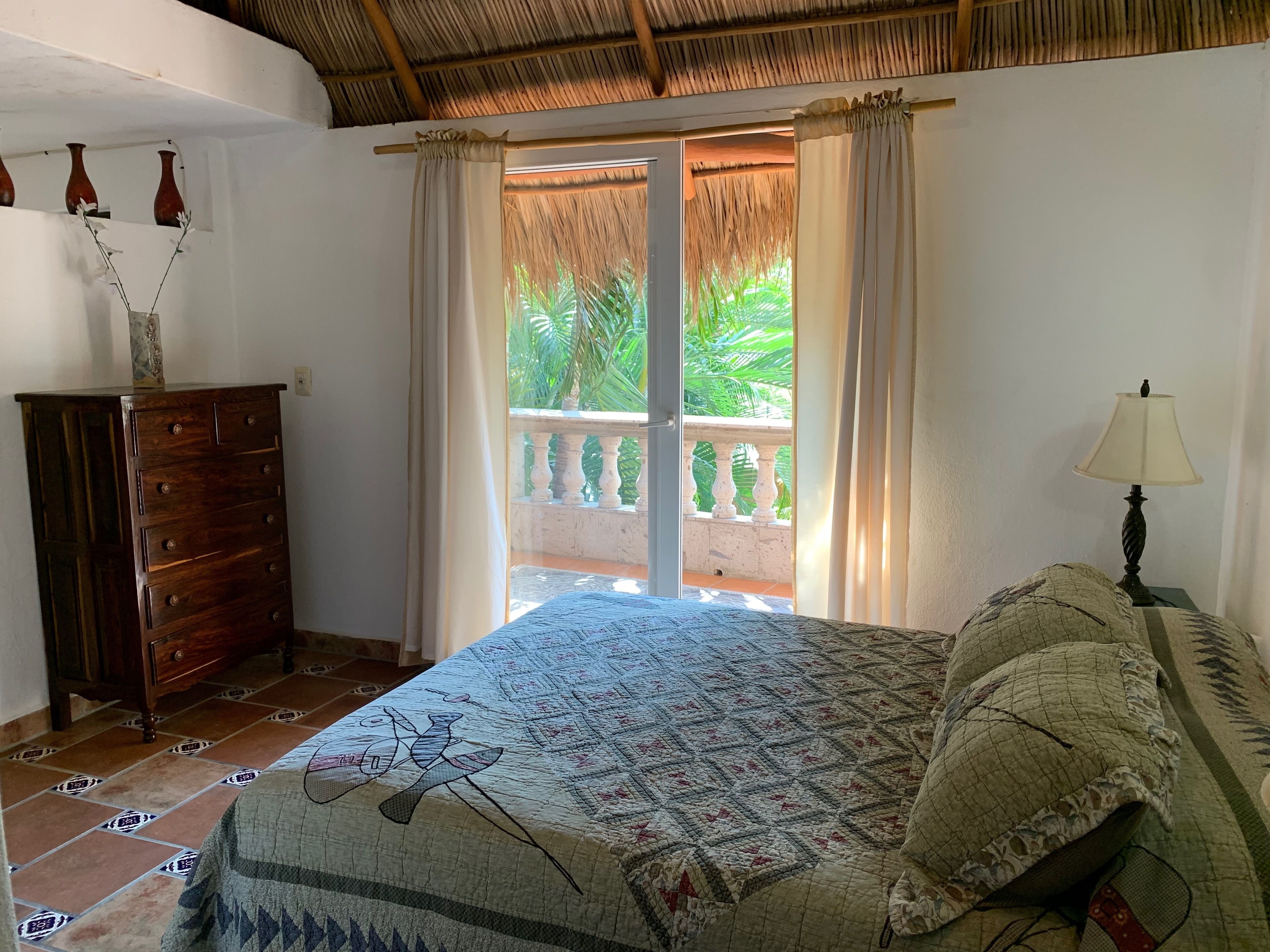 A bedroom with a bed, a dresser, a lamp, and a view of a balcony with plants.